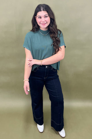 Woman wearing a green top and black jeans against a beige background