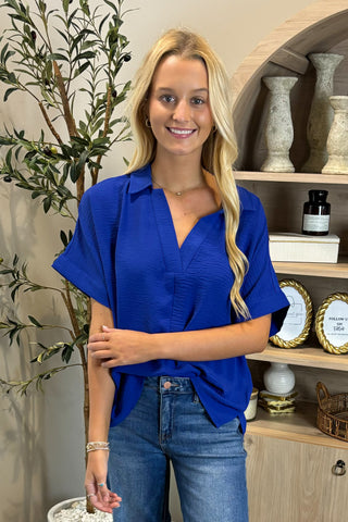 Woman wearing a blue shirt and jeans standing in front of a shelf with decorative items.