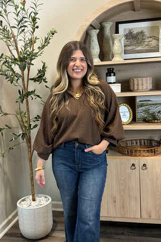 Woman in a brown sweater and blue jeans standing in a room with a plant and shelves.
