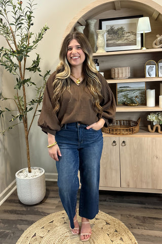 Woman in brown sweater and blue jeans standing in a room with a plant and shelves.