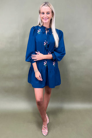 Woman wearing a blue dress with floral embroidery against a plain background