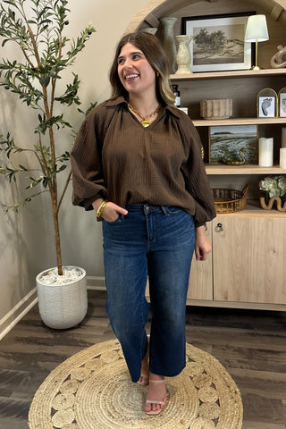 Woman in a brown blouse and blue jeans standing in a room with a plant and shelves.