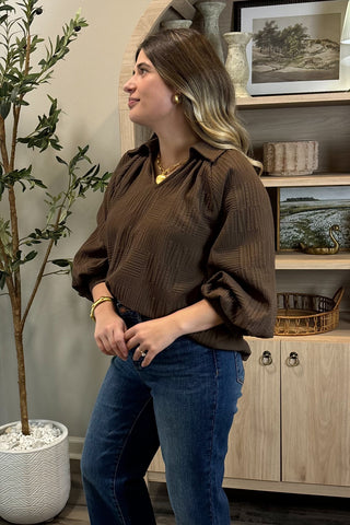 Woman in a brown blouse and blue jeans standing in a room with a plant and shelves.