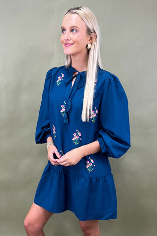 Woman wearing a blue dress with floral embroidery against a plain background