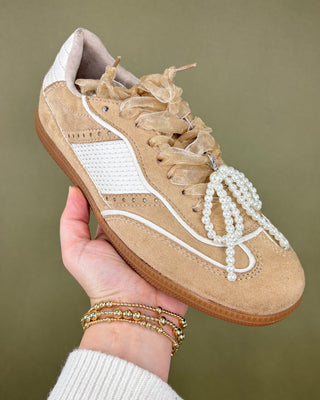 Beige sneaker with pearl laces held by a hand against a green background