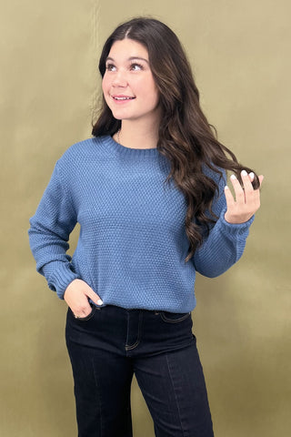 Woman wearing a blue sweater and black pants against a beige background