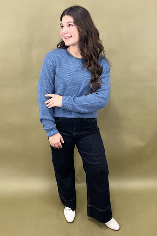 Woman wearing a blue sweater and black pants against a beige background