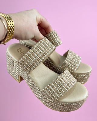 Beige platform sandals with rhinestone straps held by a hand against a pink background