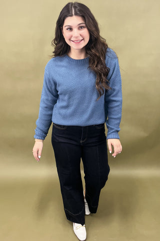 Woman wearing a blue sweater and black pants against a beige background