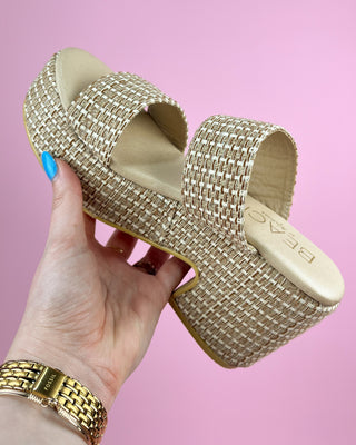 Gold woven sandals held by a hand with a gold bracelet against a pink background