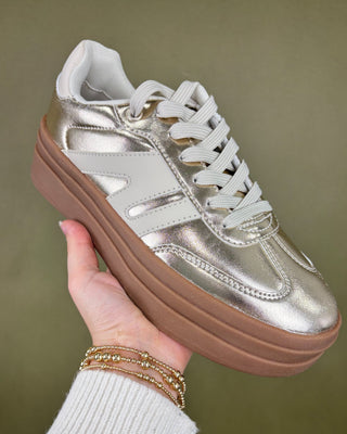 gold sneakers with brown sole held by a hand against a green background