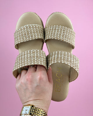 Gold embellished sandals with 'Beach' text held by a hand against a pink background