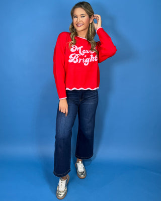 Red “Merry + Bright” Christmas sweater with pink lettering, scalloped pink trim, crew neckline, and long sleeves.