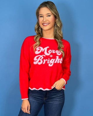 Red “Merry + Bright” Christmas sweater with pink lettering, scalloped pink trim, crew neckline, and long sleeves.