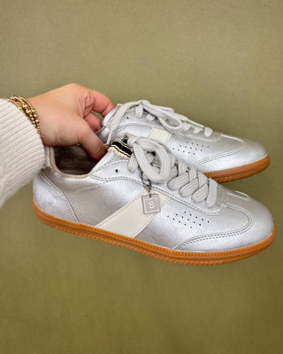Silver sneakers with brown soles held by a hand against a beige background