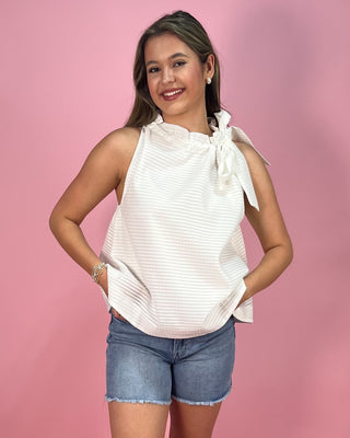 Woman wearing a white sleeveless top with a bow and denim shorts on a pink background