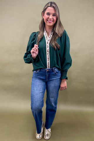 Woman wearing a green blouse with lace details and blue jeans against a beige background