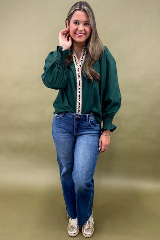Woman wearing a green blouse with lace details and blue jeans against a beige background