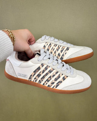 White sneakers with brown soles held by a hand against a beige background