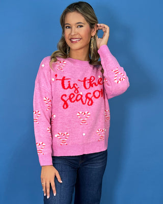 Pink “’Tis the Season” Christmas sweater with candy cane hearts, red lettering, white pom-pom accents, and long sleeves.