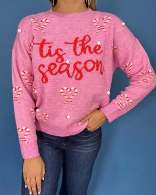 Pink “’Tis the Season” Christmas sweater with candy cane hearts, red lettering, white pom-pom accents, and long sleeves.