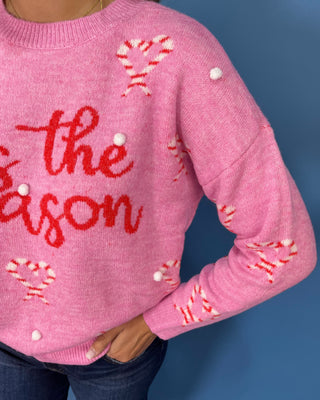 Pink “’Tis the Season” Christmas sweater with candy cane hearts, red lettering, white pom-pom accents, and long sleeves.