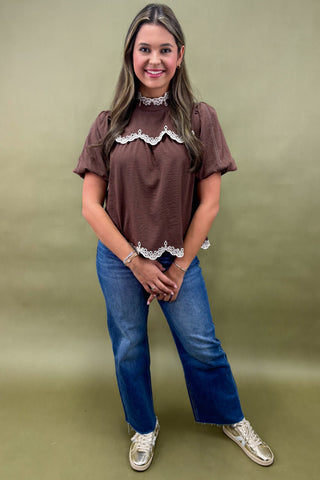 Woman wearing a brown top with lace details and blue jeans on a green background