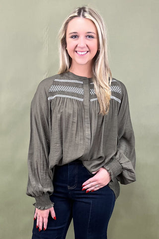 Woman wearing an olive green blouse with white embroidery against a plain background