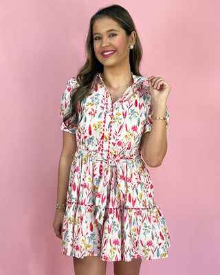 Model wearing a white floral mini dress with puff sleeves, a button-front bodice, and a self-tie waist, finished with a flouncy tiered skirt and styled with white heeled sandals against a pink backdrop.