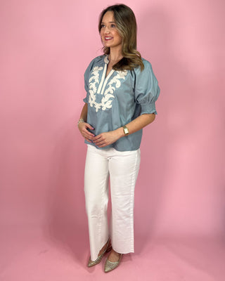 Woman wearing a blue embroidered blouse and white pants against a pink background