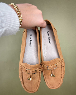 Pair of tan loafers with 'Pierre Dumas' branding held by a hand against a green background