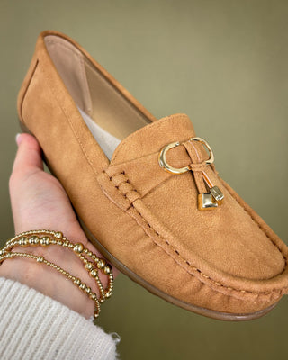 Tan loafer with gold buckle held by a hand against a green background