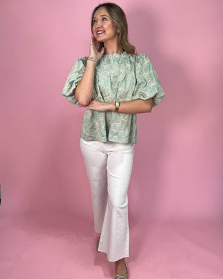 Woman wearing a green patterned top and white pants against a pink background