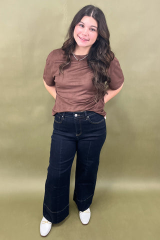 Woman wearing a brown top and dark blue jeans against a green background