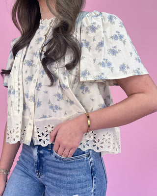 Woman wearing a floral blouse with eyelet details and blue jeans against a pink background