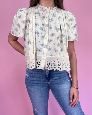 Person wearing a floral blouse with lace details and blue jeans on a pink background