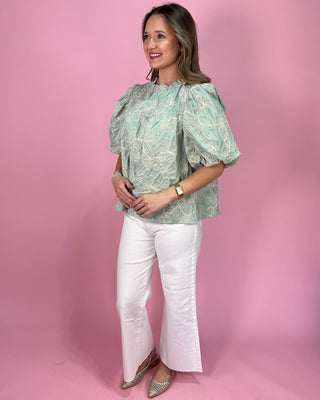 Woman wearing a green floral blouse and white pants against a pink background