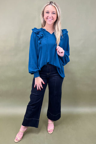 Woman wearing a blue blouse with ruffles and black pants on a beige background