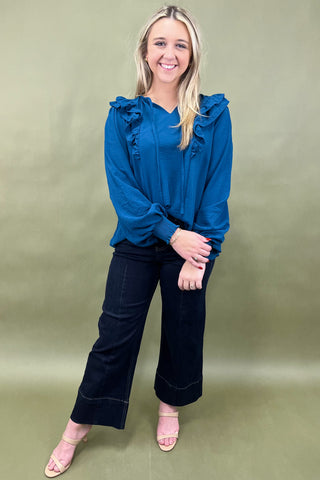 Woman wearing a blue blouse with ruffles and dark pants on a green background
