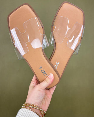 Clear strap flat shoes held by a hand against a green background