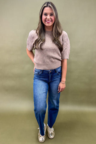 Woman wearing a beige sweater and blue jeans against a green background