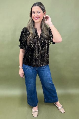 Woman wearing a black patterned blouse and blue jeans against a green background