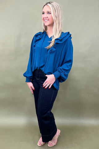 Woman wearing a blue blouse with ruffles and black pants against a plain background