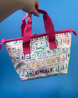 A hand holds a Swig brand tote bag with a white background and colorful word tiles, featuring bright pink handles and trim against a solid blue background.