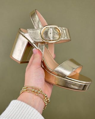 Gold high-heeled sandal held by a hand against a plain background