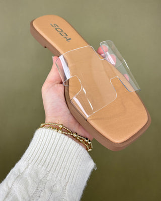 Hand holding a tan sandal with clear plastic insole against a green background