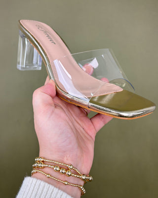 Gold high-heeled shoe with clear heel held by a hand against a green background