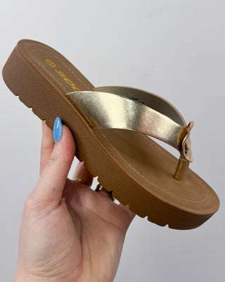 Gold sandal with brown sole held by a hand against a gray background