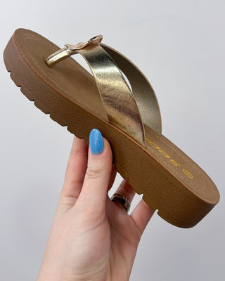 Brown sandal with gold strap held by a hand against a gray background