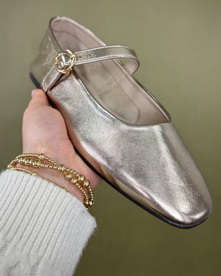 Metallic shoe held by a hand with gold bracelets on a neutral background